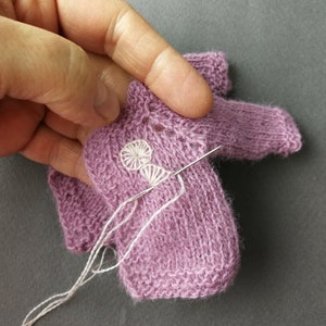 May include: A close-up of a purple knitted sweater with a white embroidered flower design. A needle and white thread are being used to stitch the flower.
