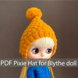 Può includere: Un cappello da elfo di lana gialla con un pon pon in cima. Il cappello ha lunghi lacci che possono essere legati sotto il mento. Il cappello è indossato da una bambola con i capelli biondi e grandi occhi blu. Il testo "PDF Pixie Hat for Blythe doll" è in fondo all'immagine.