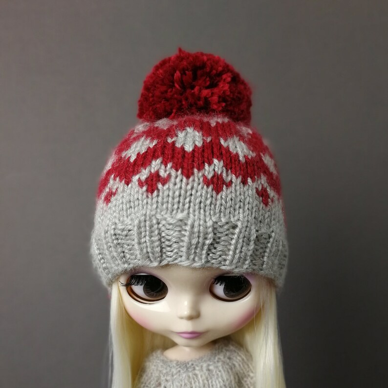 May include: A doll wearing a gray and red knitted hat with a red pom-pom on top. The hat has a fair isle pattern.