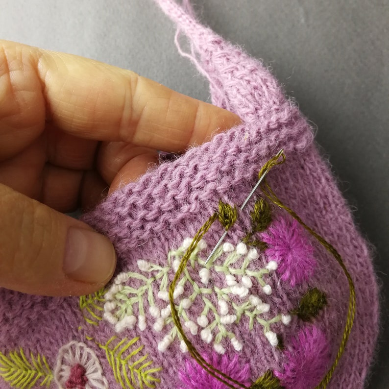 May include: Close-up of a purple knitted fabric with embroidered flowers in white, green, and pink. A needle and thread are visible, suggesting the embroidery is in progress.