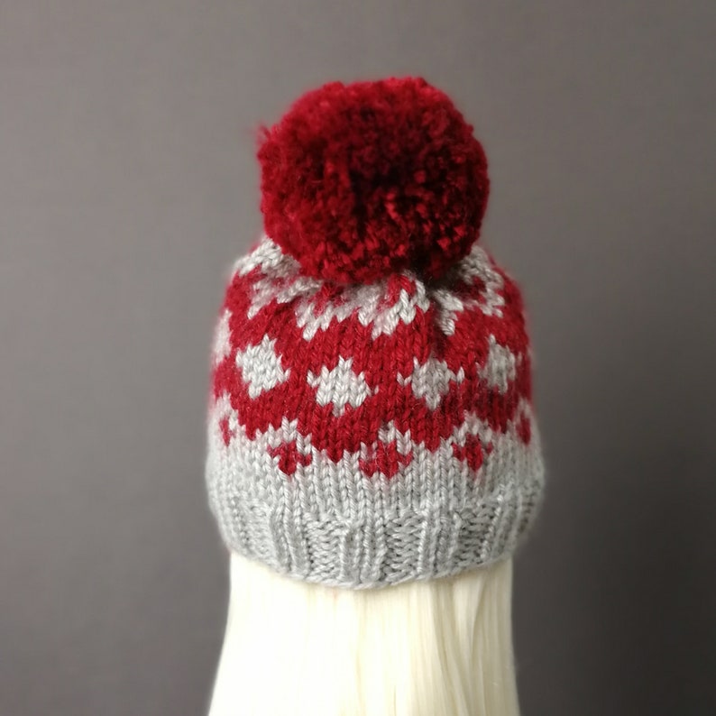 May include: A red and gray knitted hat with a pom pom on top. The hat has a fair isle pattern with white snowflakes on a red background.