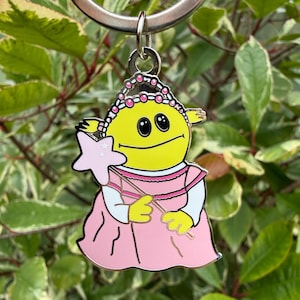 Könnte beinhalten: Ein Schlüsselanhänger aus Metallemaille mit einer gelben Zeichentrickfigur mit rundem Kopf, schwarzen Augen und einem dünnen Lächeln. Die Figur trägt ein rosa Kleid mit weißen Ärmeln und ein rosa Diadem und hält einen hellrosa sternförmigen Zauberstab.