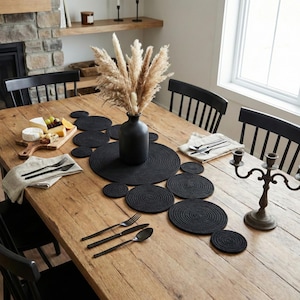 Könnte beinhalten: Ein rustikaler Holzesstisch, der für eine Mahlzeit gedeckt ist. Der Tisch ist mit schwarzen Tischsets, Untersetzern und einem Mittelstück aus getrocknetem Pampasgras in einer schwarzen Vase geschmückt. Besteck und ein Käsebrett sind ebenfalls vorhanden.