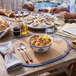 Coastal blue jute placemats on a bright table with plates, glassware and green foliage near a window.
