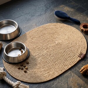 Oval jute dog bowl mat on a dark slate floor, holding two stainless steel pet bowls with a rope toy beside it.