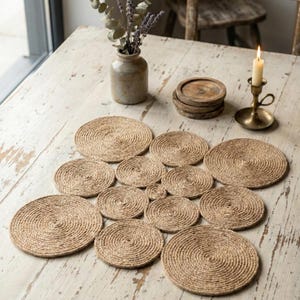 Pode incluir: Um conjunto de bases para copos circulares de juta tecida dispostas sobre uma mesa de madeira branca desgastada. Um pequeno vaso com flores secas, um castiçal de latão e bases para copos de madeira também estão presentes.