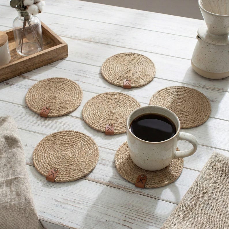 Six jute rope coasters with faux leather tags on whitewashed planks, with a wooden tray holding cotton stems in a glass vase, a stoneware pour over jug and a speckled mug of black coffee on linen