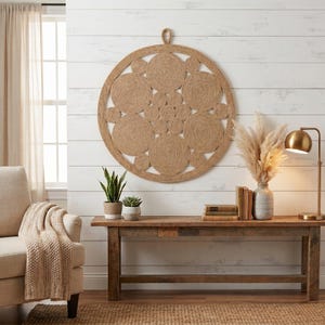 Round jute wall art on white shiplap above a rustic console table with pampas grass, a brass lamp, antique books and a cream armchair