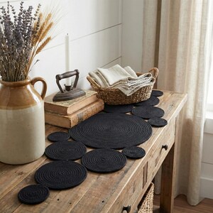 May include: Vintage farmhouse console table styled with a handmade black cotton runner featuring interconnected round braided patterns. The rustic home decor includes a ceramic vase with dried lavender, a woven basket, and an antique iron for a charming entryway look.