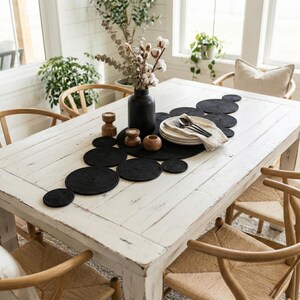 May include: Whitewashed rustic wooden dining table adorned with a modern black cotton woven runner. The striking round geometric design adds a boho touch, beautifully contrasted with natural wood chairs, a black vase, and simple tableware in a bright, airy room.