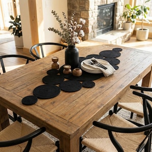 May include: A rustic wooden dining table set with black placemats, coasters, and a centerpiece. The table is surrounded by black chairs with woven seats. A black vase holds cotton stems and eucalyptus.