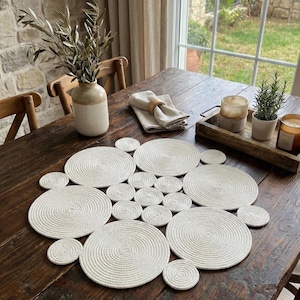 May include: A wooden dining table displays a set of white, circular placemats and coasters. A vase with olive branches, a linen napkin, and a wooden tray with candles and a plant are also present. The table is near a window, creating a warm, inviting atmosphere.