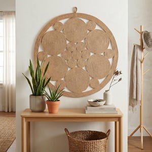 Large jute wall art above a light oak console with snake plants, a ceramic vase of dried stems, books and a woven basket beside a wooden coat stand