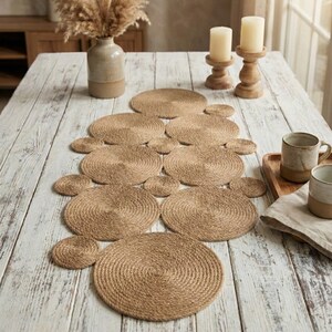 May include: A table setting with woven jute placemats and coasters. The placemats are arranged in a line down the centre of a light-coloured wooden table. Two ceramic mugs and candles in wooden holders complete the natural, neutral aesthetic, creating a rustic look.