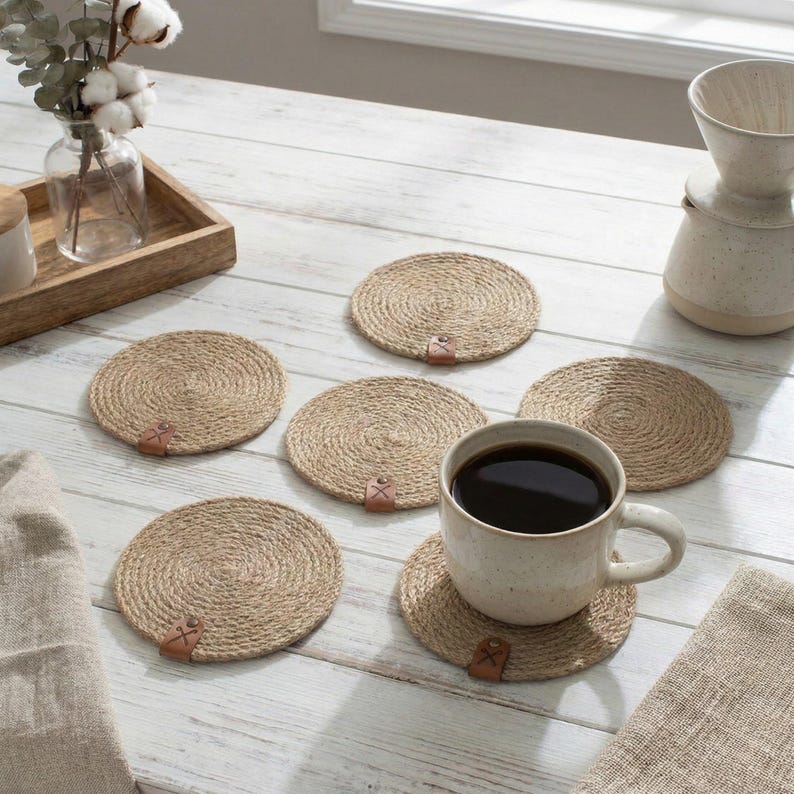 Six jute rope coasters with faux leather tags on a whitewashed table by a window, with cotton and eucalyptus in a glass vase on a wooden tray, a stoneware pour over jug and a speckled mug of coffee