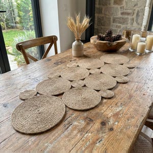 May include: A natural jute table runner made of circular placemats. The placemats are arranged in a connected pattern on a rustic wooden table. The table is decorated with a vase of wheat and other decorative items.