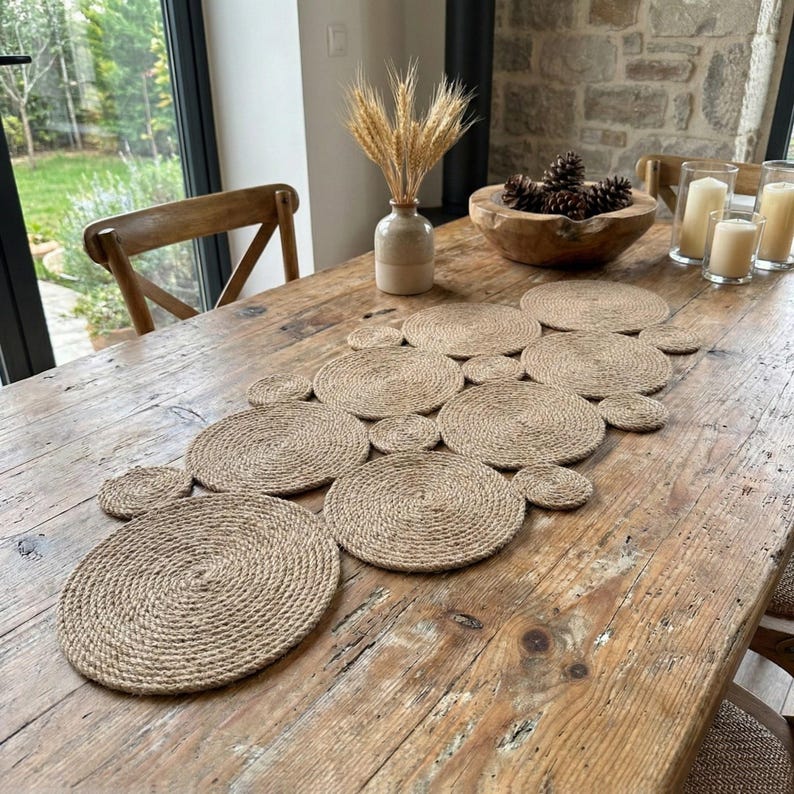 May include: A natural jute table runner made of circular placemats. The placemats are arranged in a connected pattern on a rustic wooden table. The table is decorated with a vase of wheat and other decorative items.