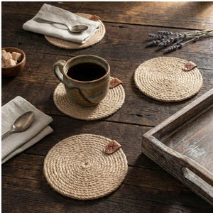 Four round jute coasters with faux leather tags on dark reclaimed wood, with a reactive glaze mug of coffee, a bowl of brown sugar cubes, lavender sprigs, linen napkins with silver spoons and a weathered wooden tray