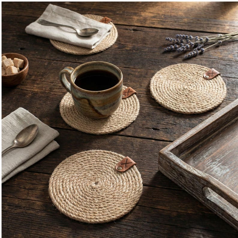 Four round jute coasters with faux leather tags on dark reclaimed wood, with a reactive glaze mug of coffee, a bowl of brown sugar cubes, lavender sprigs, linen napkins with silver spoons and a weathered wooden tray
