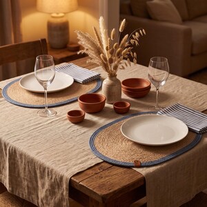 Two oval jute placemats with a blue stripe border on a linen tablecloth, set with white plates, wine glasses, terracotta bowls and dried grasses in a stoneware vase under warm lamplight