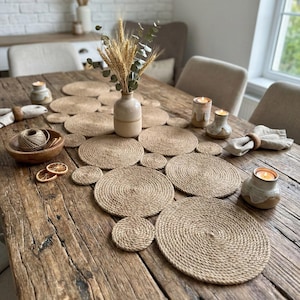 Pode incluir: Uma mesa rústica de madeira é decorada com jogos americanos e porta-copos de juta tecida. Um vaso bege contém trigo e eucalipto. Velas e uma tigela de madeira com barbante completam a decoração natural.