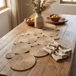 May include: A natural-toned dining table setting with a jute table runner and circular placemats. Linen napkins with wooden ring holders are arranged alongside. A vase with dried flowers and wooden bowls with fruit complete the rustic decor.