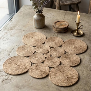 May include: A selection of circular jute placemats, arranged on a concrete surface. The placemats are made from natural-coloured jute rope and vary in size. A vase with dried flowers, wooden coasters, and a candle in a brass holder are also on the table.