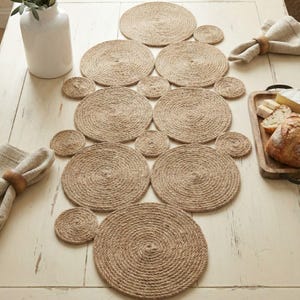 May include: A set of woven jute placemats and coasters on a light wooden table. The circular placemats and coasters are made from tightly coiled natural jute rope. Napkins with wooden rings and a vase are also visible.
