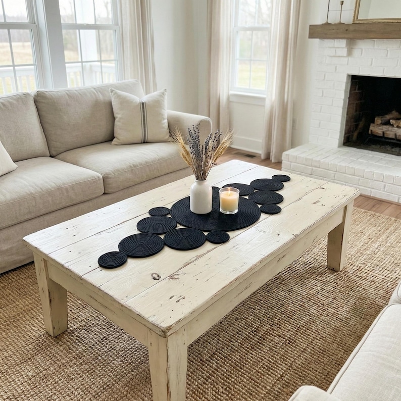 May include: Minimalist and bright living room decor showcasing a handcrafted black cotton table runner on a whitewashed wooden coffee table. The unique interconnected circular design provides a bold contrast against the neutral tones of the white sofa and pampas grass.