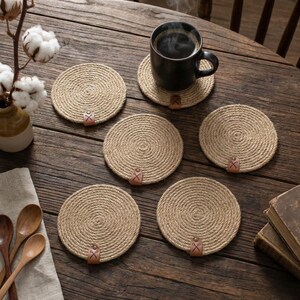 Six round jute coasters with faux leather tags on a dark weathered table, with cotton stems in an amber stoneware jug, a steaming black mug, wooden spoons on a linen napkin and stacked antique books