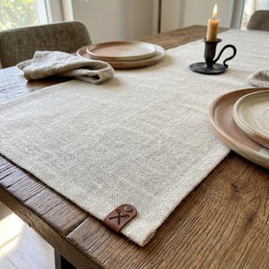 May include: A neutral-toned linen table runner with a brown leather tag featuring a scissors and fork design. The runner is placed on a wooden table, with plates, a candle, and a napkin visible. The overall theme is rustic dining.