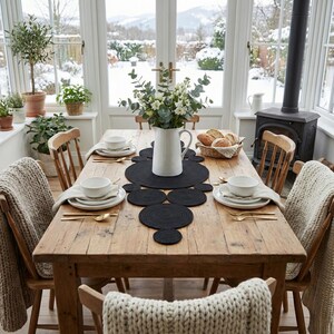 May include:Cozy winter farmhouse dining table decorated with a striking black cotton table runner. The braided circular centerpiece creates an inviting atmosphere, complemented by white tableware, a white pitcher vase with fresh greenery, and a snowy window view.