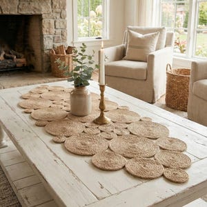 Könnte beinhalten: Rustikaler Tischläufer auf einem Couchtisch, bestehend aus gewebten Jute-Tischsets, die in einem kreisförmigen Muster angeordnet sind. Eine brennende Kerze in einem Messinghalter, eine Vase mit Grün und ein weißer Tisch.
