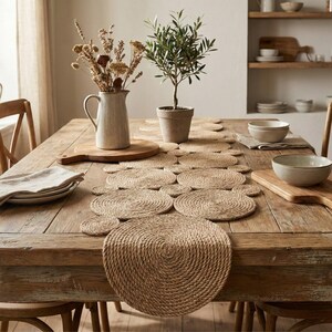 May include: A wooden dining table features a jute table runner and circular placemats. A ceramic jug with dried flowers, a small potted olive tree, and serving bowls create a natural, neutral-toned aesthetic. The table is set for dining.
