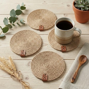 Four coiled jute coasters with faux leather tags on pale pine planks, with a eucalyptus sprig, a succulent in a terracotta pot, a bundle of wheat and a speckled mug of coffee beside a wooden spoon on linen
