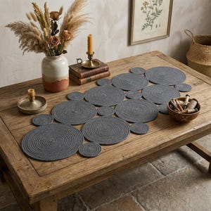 Könnte beinhalten: Ein Satz grauer runder Tischsets auf einem Holztisch. Die Tischsets bestehen aus gewebtem Material und sind in verschiedenen Größen erhältlich. Der Tisch ist von dekorativen Gegenständen umgeben, darunter eine Vase mit Trockenblumen, Kerzen und Büchern.