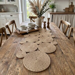May include: A dining table made of weathered wood is set with a woven jute placemat and coasters. Wooden chairs surround the table. A vase of dried flowers and greenery sits in the centre of the table.