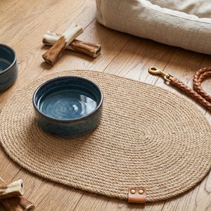 Handmade 100% natural jute dog bowl mat on a rustic light wood floor. This fringeless, hand-stitched placemat features a premium genuine leather tag and serves as a stylish, durable feeding station for dogs. The organic woven texture protects floors from food and water spills while providing a rustic, farmhouse aesthetic that blends seamlessly with modern pet home decor. Perfect for sustainable pet owners looking for functional and aesthetic dining solutions for their furry friends.