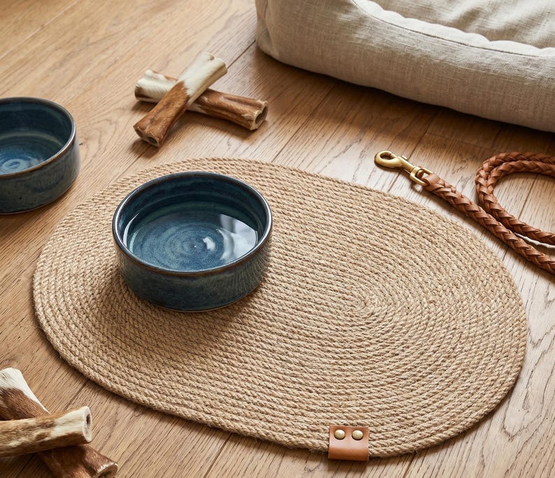 Handmade 100% natural jute dog bowl mat on a rustic light wood floor. This fringeless, hand-stitched placemat features a premium genuine leather tag and serves as a stylish, durable feeding station for dogs. The organic woven texture protects floors from food and water spills while providing a rustic, farmhouse aesthetic that blends seamlessly with modern pet home decor. Perfect for sustainable pet owners looking for functional and aesthetic dining solutions for their furry friends.