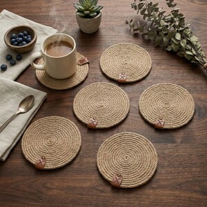 Six coiled jute coasters with faux leather tags on a dark walnut table, with a milky coffee in a cream mug, a bowl of blueberries, a succulent in a grey pot, a eucalyptus branch and a silver spoon on linen