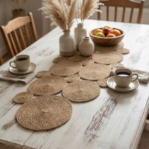 May include: A dining table set with a jute placemat and coasters. Two cups of coffee with saucers, a wooden fruit bowl, and vases with dried plants are on the table. The table is painted white with a distressed finish.