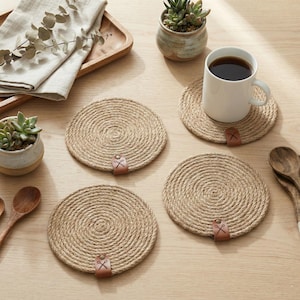Four coiled jute coasters with faux leather tags on a pale oak table, with a white mug of coffee, two succulents in speckled pots, a wooden tray with linen and eucalyptus, and wooden spoons