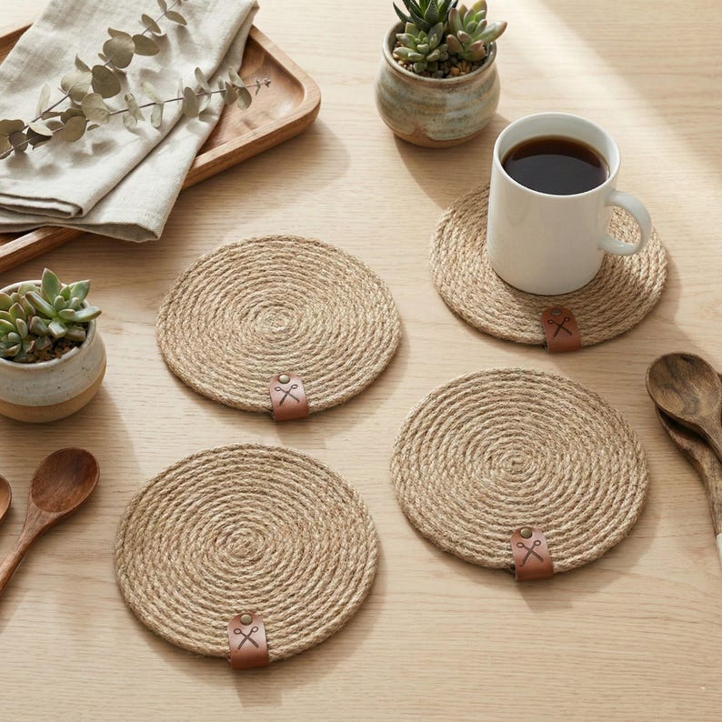 Four coiled jute coasters with faux leather tags on a pale oak table, with a white mug of coffee, two succulents in speckled pots, a wooden tray with linen and eucalyptus, and wooden spoons
