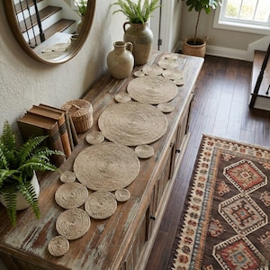 Weathered console table in an entryway styled with a jute disc runner, stacked antique books, a fern and a stoneware pitcher, with a patterned kilim rug on the floor