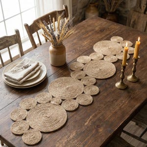 Könnte beinhalten: Ein rustikaler Esstisch mit einem Tischläufer aus geflochtenem Jute mit kreisförmigen Designs. Der Tisch ist mit Tellern, Leinen-Servietten, einer Vase mit Trockenblumen und brennenden Kerzen in Messinghaltern gedeckt. Der Holztisch hat einen warmen, einladenden Ton.