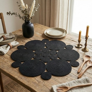 May include: A wooden table displays a black rope placemat and coasters. The table is set with plates, napkins, wooden utensils, a black vase with dried flowers, and brass candle holders with lit candles.