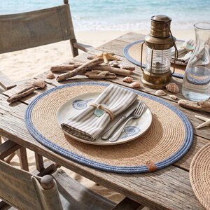 Denim blue striped jute placemats on a coastal table with sea view, styled with white plates and napkins.