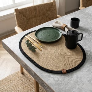 May include: A dining table setting featuring a woven jute placemat with a black border. A green plate, gold cutlery, a black teapot, and a black cup are arranged on the placemat. A woven chair is in the background.