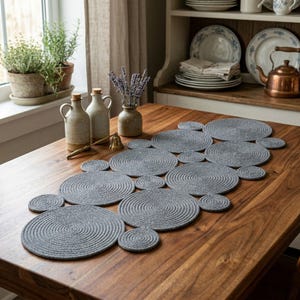 May include: A gray, circular rope table runner with multiple connected circles, placed on a wooden table. The runner is surrounded by decorative items, including vases, a bell, and a window with plants.
