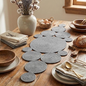 Könnte beinhalten: Ein rustikaler Holztisch ist mit einem grauen Platzset und Untersetzern aus Seil ausgestattet. Eine Vase mit Blumen, ein Korb mit Eiern und ein Brotlaib sind ebenfalls auf dem Tisch. Leinen-Servietten und Holzschalen vervollständigen die Szene.